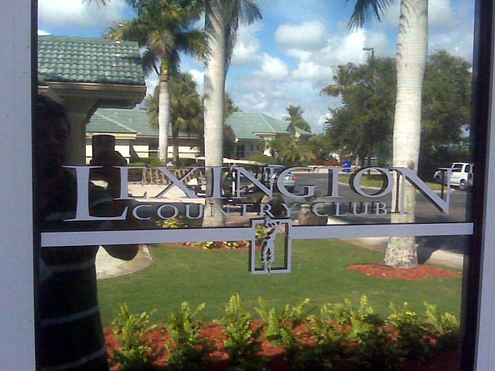 Lexington Country Club entrance with logo on glass window reflecting palm trees and building.