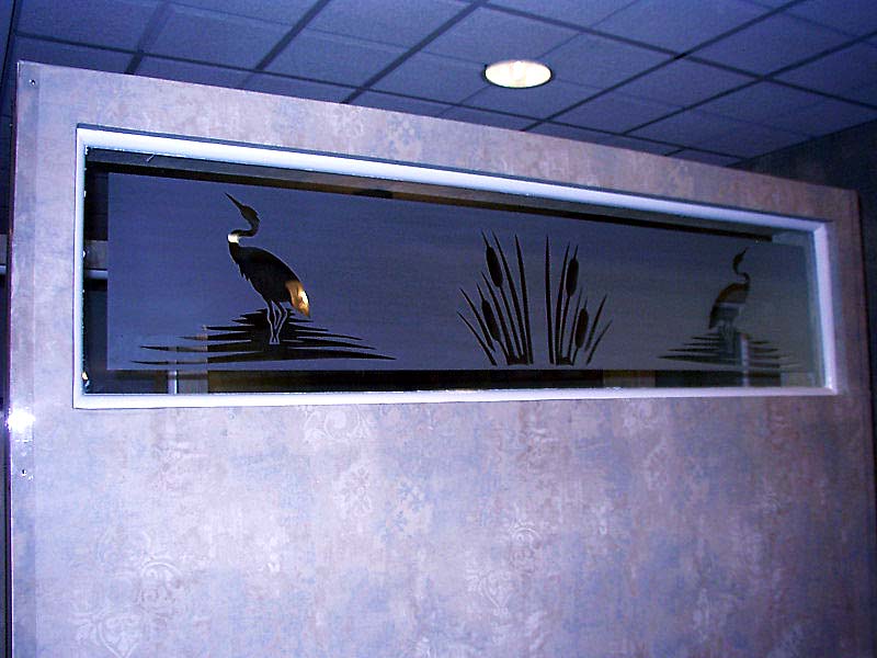 Glass window with etched silhouette of a heron, cattails, and reeds against a dark water background.