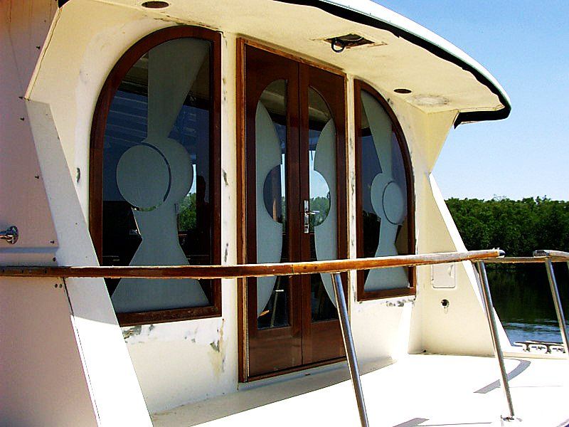 Boat exterior with rounded windows, wooden door, white paint, and railing. Sunny day with trees in the background.