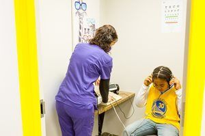 Nurse gives hearing test to child in a sound booth. Child wears headphones and a Warriors shirt. Yellow doorway.