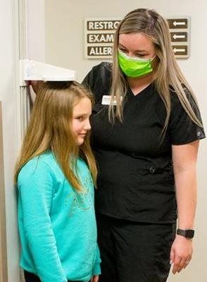 Medical professional measuring child's height against a wall. Professional wears a green face mask and black scrubs.