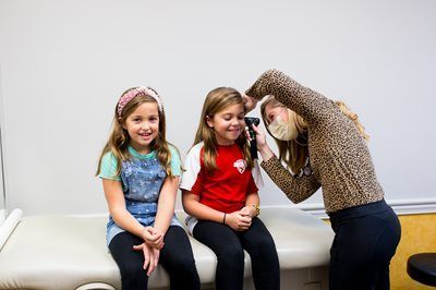 A medical professional examining a child's ear with an otoscope; two girls are sitting on a medical table, smiling.