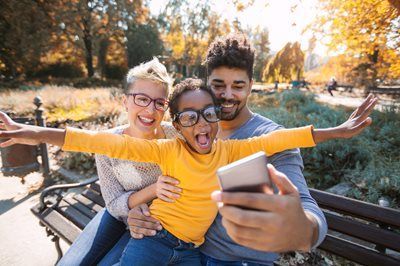 Family taking a selfie outdoors in autumn; child with arms outstretched, everyone smiling.