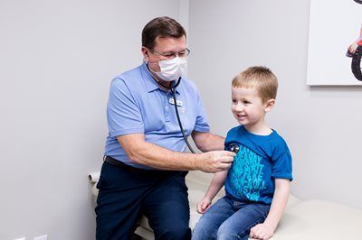 Doctor wearing mask listens to child's chest with stethoscope in a medical examination room.