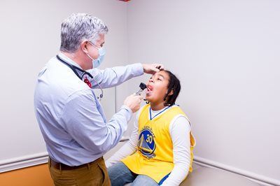 Doctor examining a person's mouth with a light. Person seated, wearing a yellow jersey. White exam room.