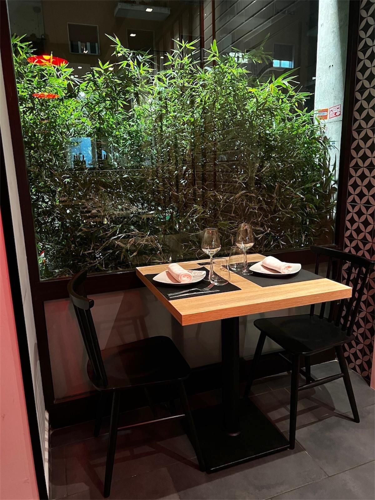 Un piccolo tavolo quadrato in legno per due, apparecchiato con piatti e bicchieri da vino, posizionato accanto a una finestra che si affaccia su una rigogliosa vegetazione.