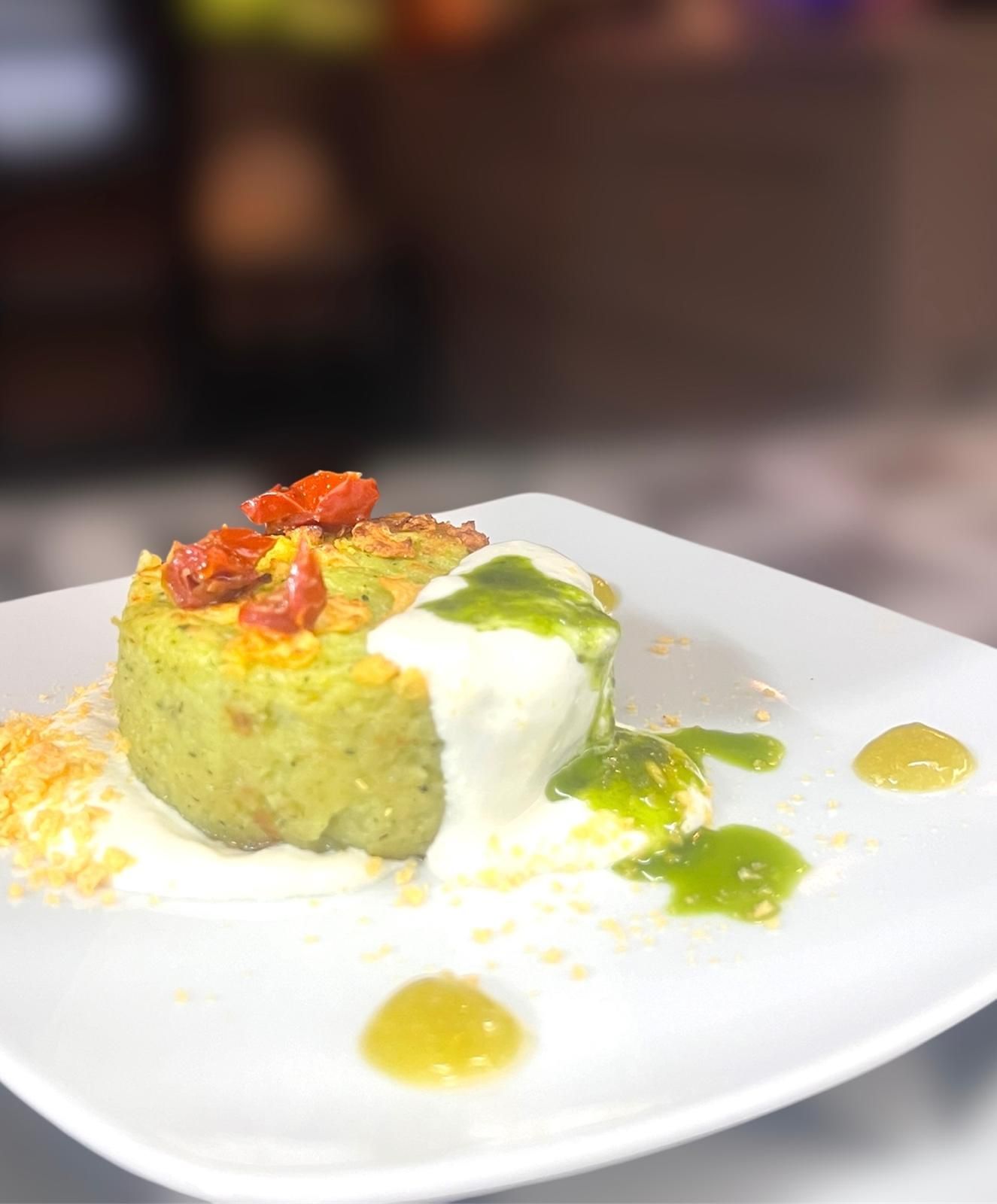 Una torta salata circolare di verdure verdi, guarnita con pomodori secchi, condita con salsa bianca e olio verde, servita su un piatto bianco.