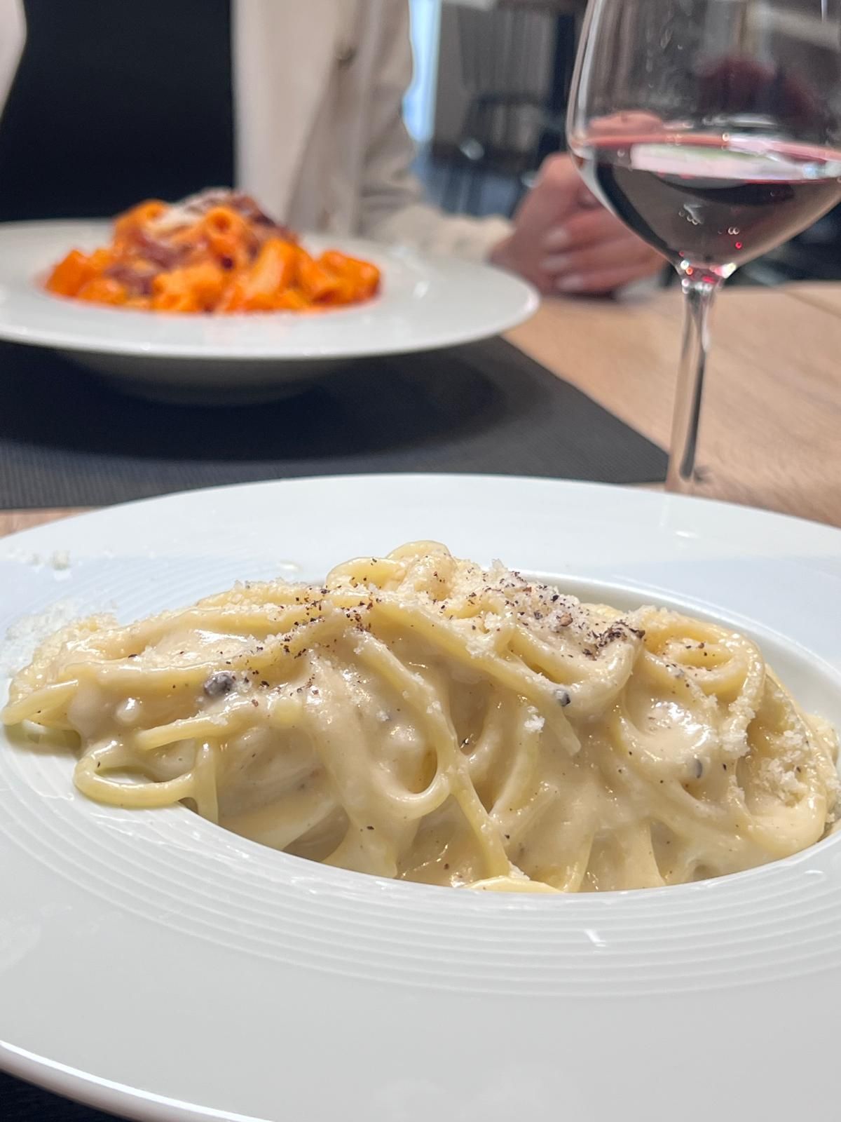 Su un tavolo, accanto a un bicchiere di vino rosso, ci sono due piatti di pasta, uno con salsa cremosa e l'altro al pomodoro.