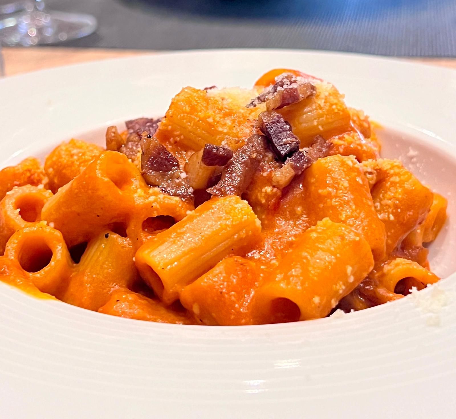 Rigatoni con una vivace salsa alla vodka arancione e pezzetti di pancetta, serviti in una ciotola bianca bassa.