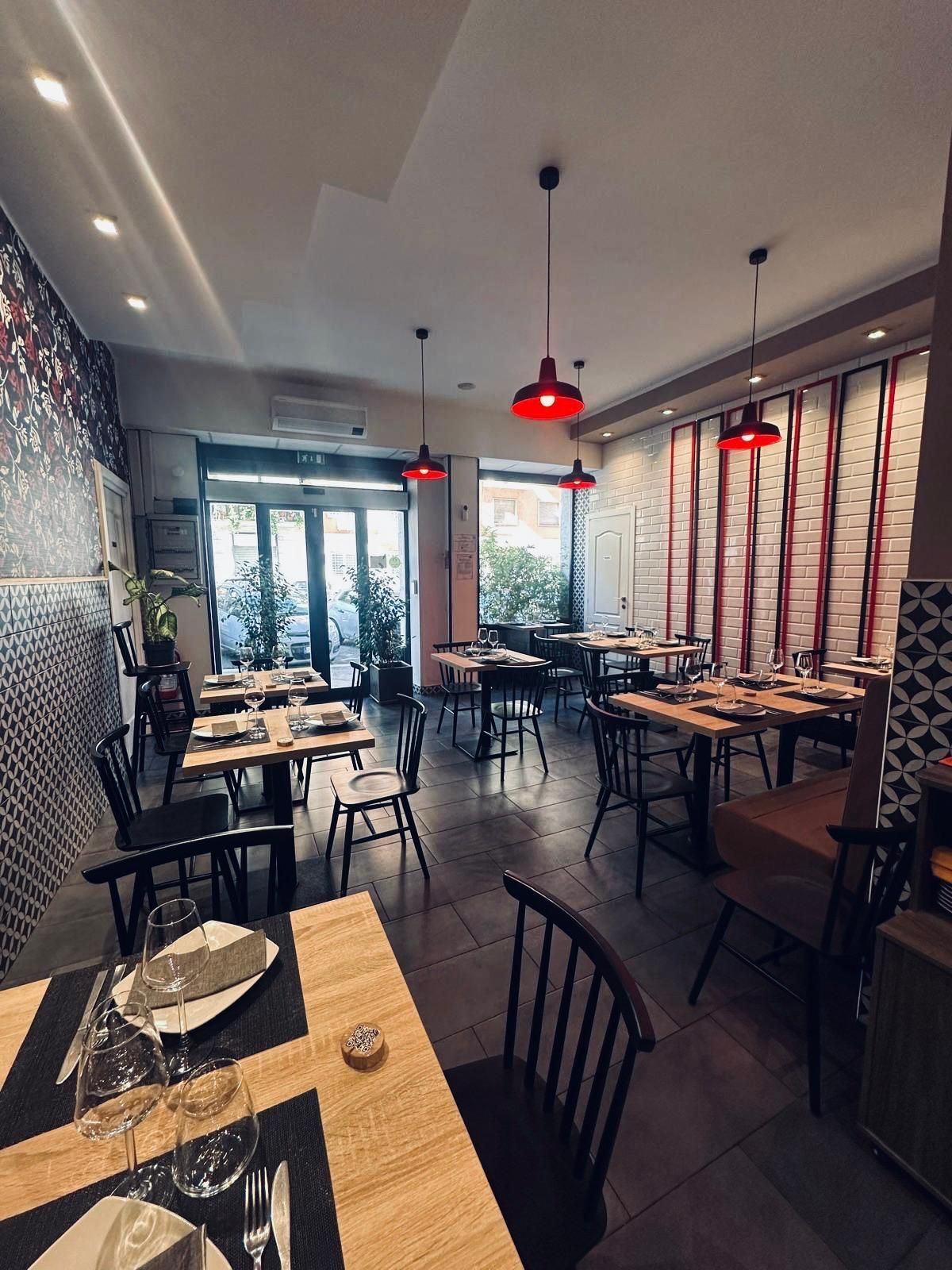 L'interno vuoto e moderno di un ristorante, con tavoli in legno, sedie nere e lampade a sospensione rosse su uno sfondo di mattoni bianchi.