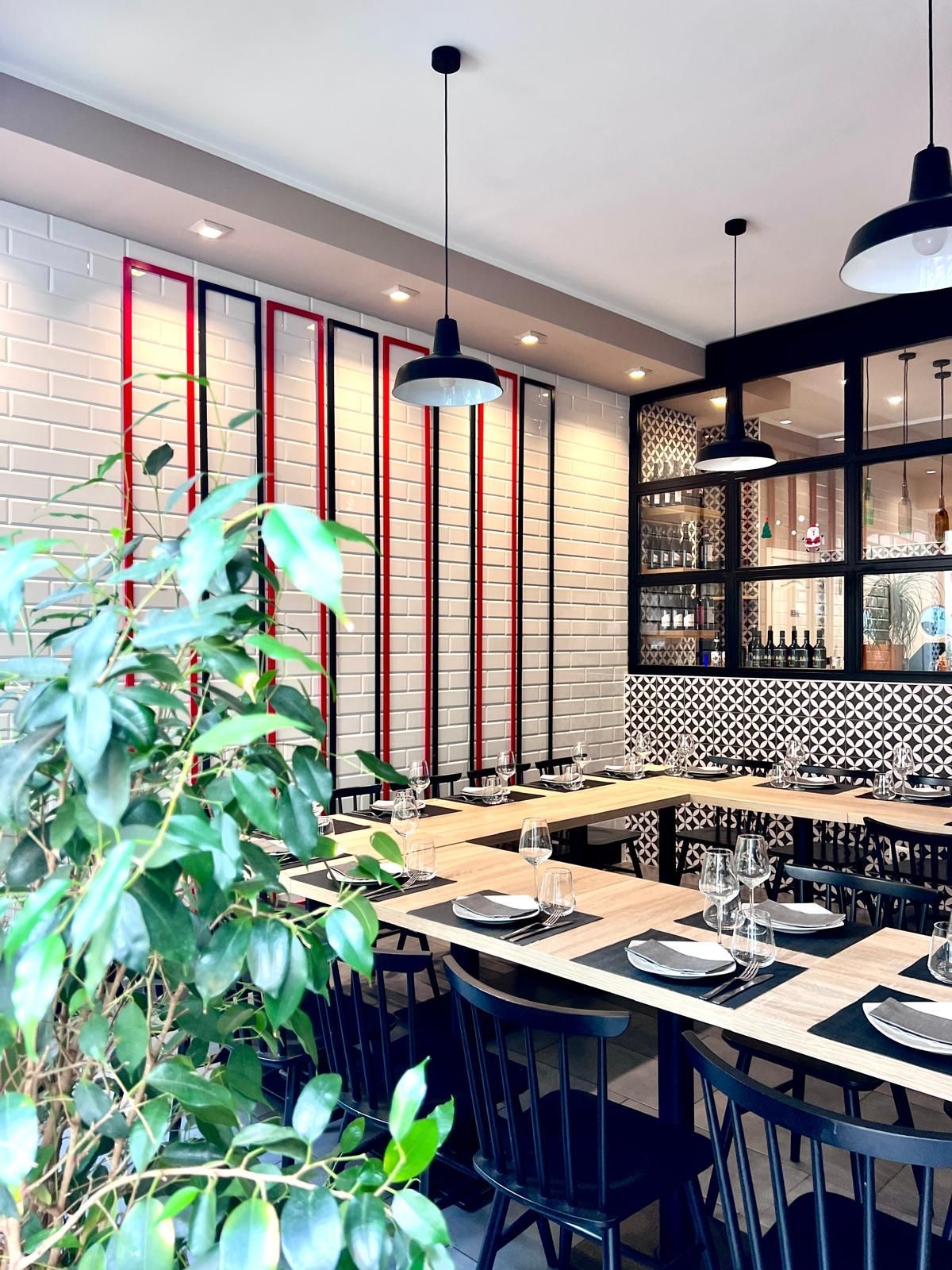 Una zona pranzo con un lungo tavolo di legno a forma di U, sedie nere con struttura a fuso, una parete di mattoni bianchi e moderne lampade a sospensione.