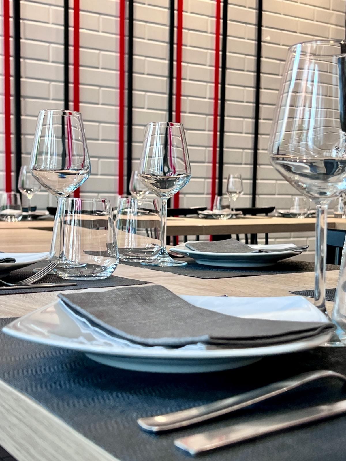 Una tavola apparecchiata con un piatto bianco, un tovagliolo grigio, posate e bicchieri da vino, con uno sfondo di muro di mattoni.