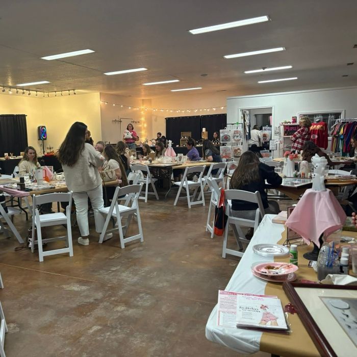 A crowded event space with tables, people, and craft displays. Warm lighting and decorations.