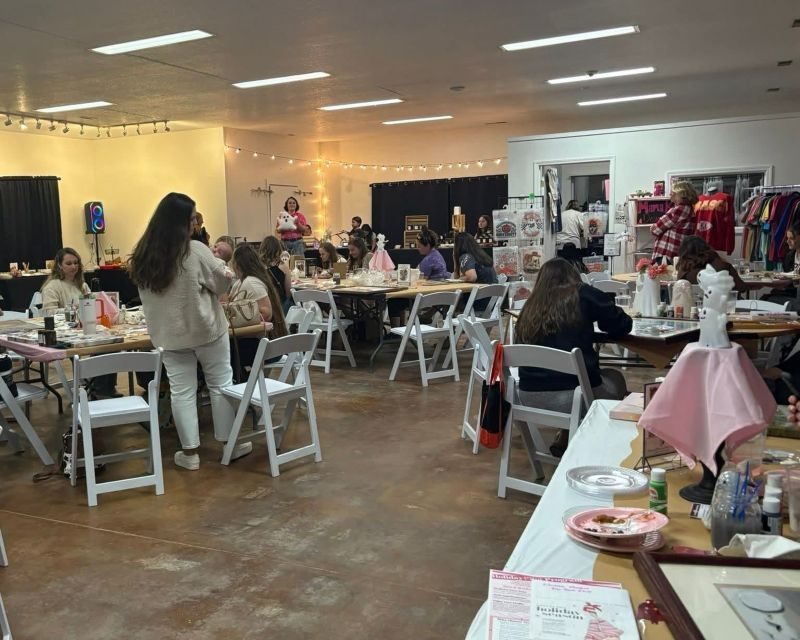 People at tables in a brightly lit room, craft fair setting. Tables are covered in items.