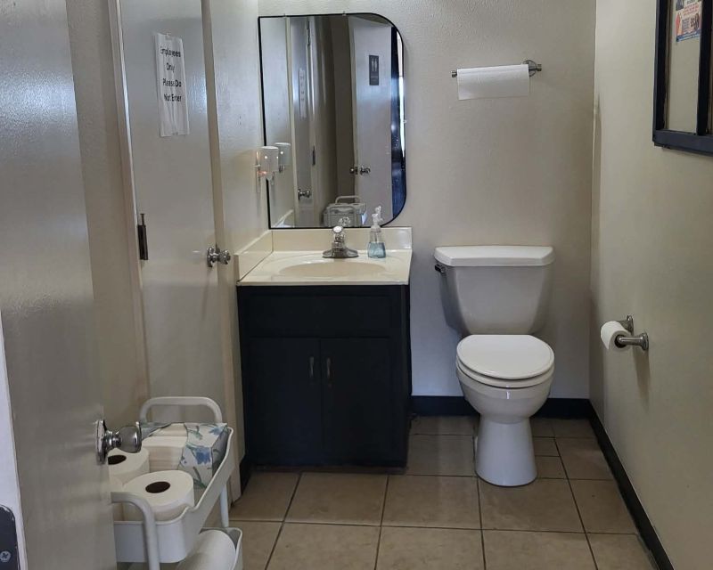 Bathroom with sink, toilet, mirror, and a small cart with paper products. Beige tile floor.