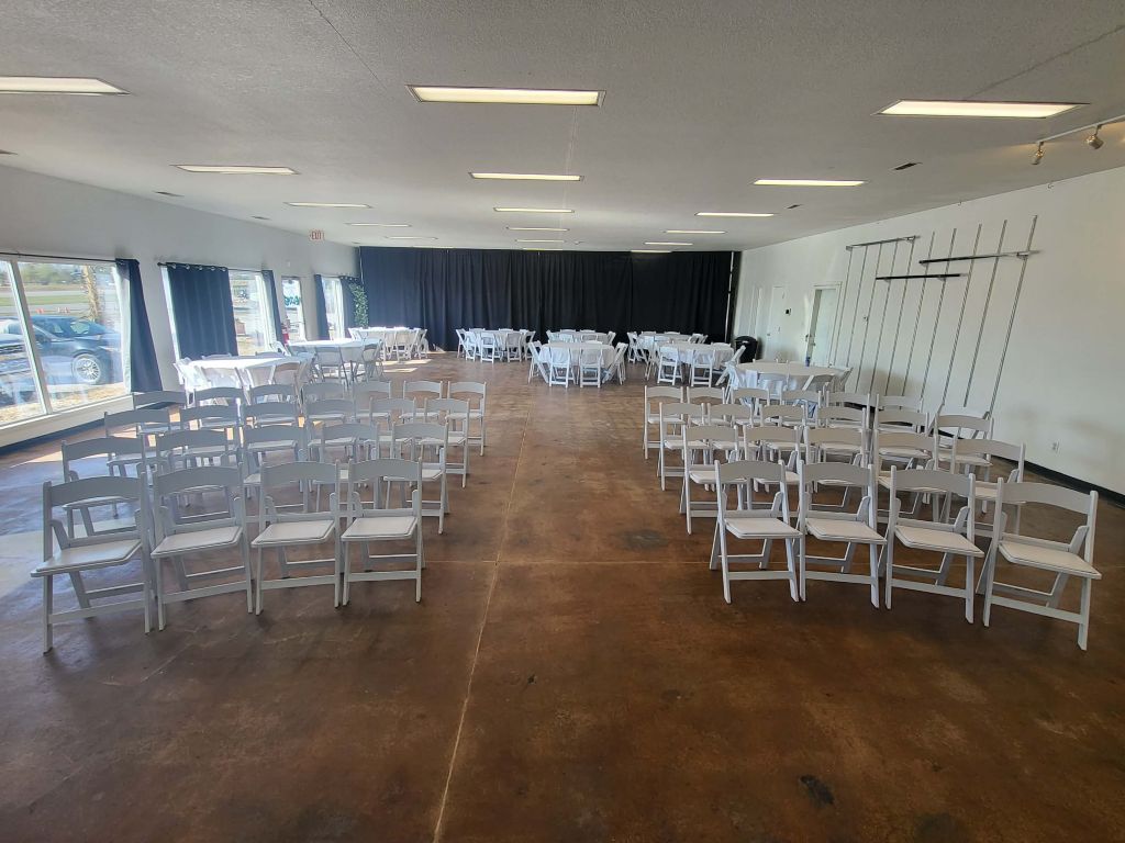 indoor event space setup with rows of white chairs facing a small stage. Tables are in the background.