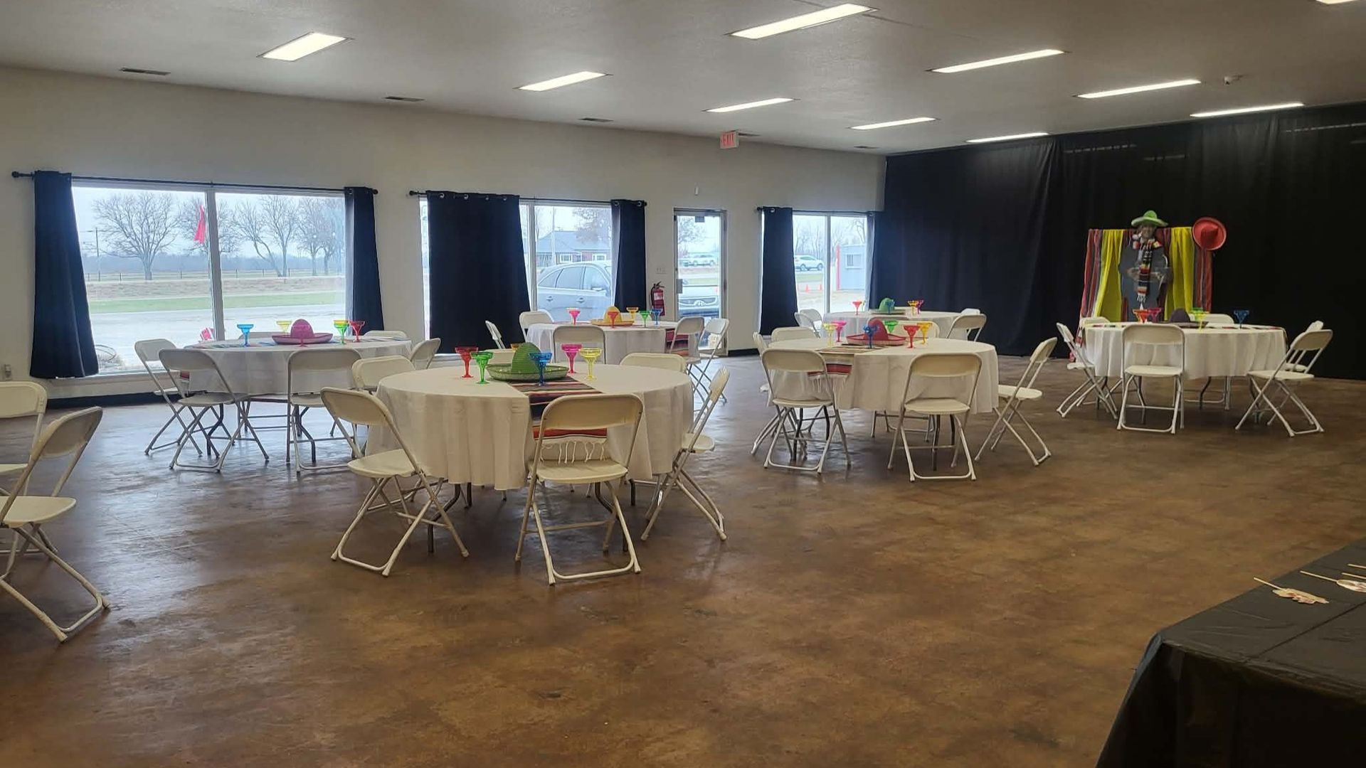 Event hall with round tables set for a party. Some tables have centerpieces; windows with black curtains.