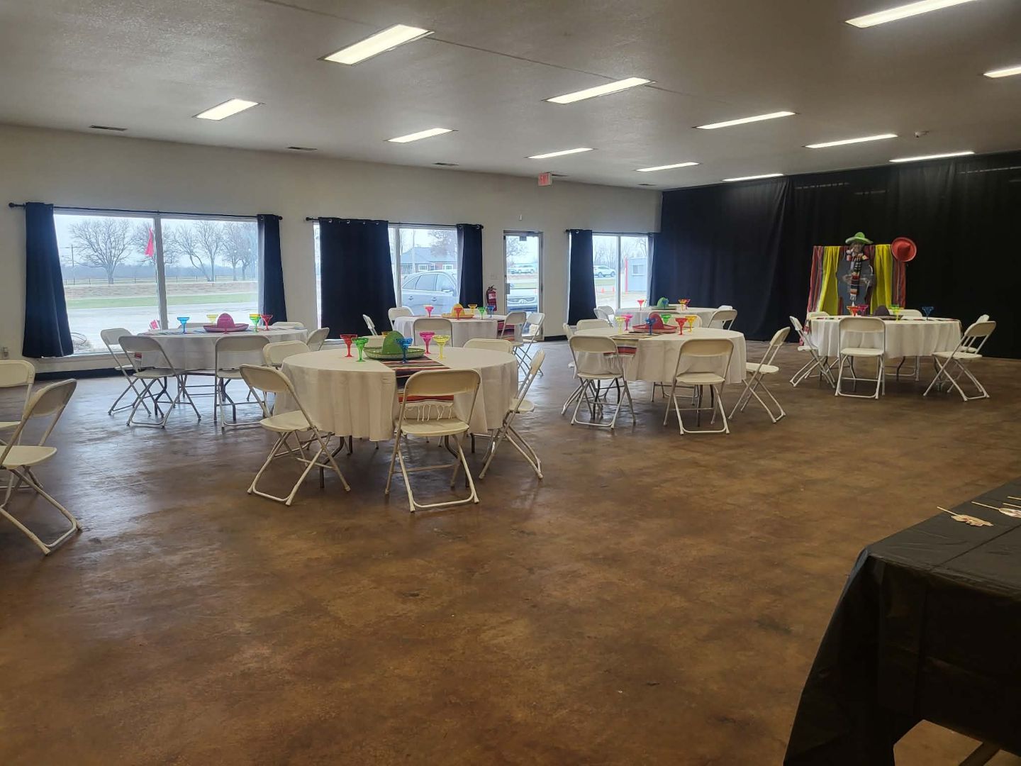 Large event room with white-clothed tables, chairs, and decorations near windows. Brown floor, dark curtains, and overhead lights.