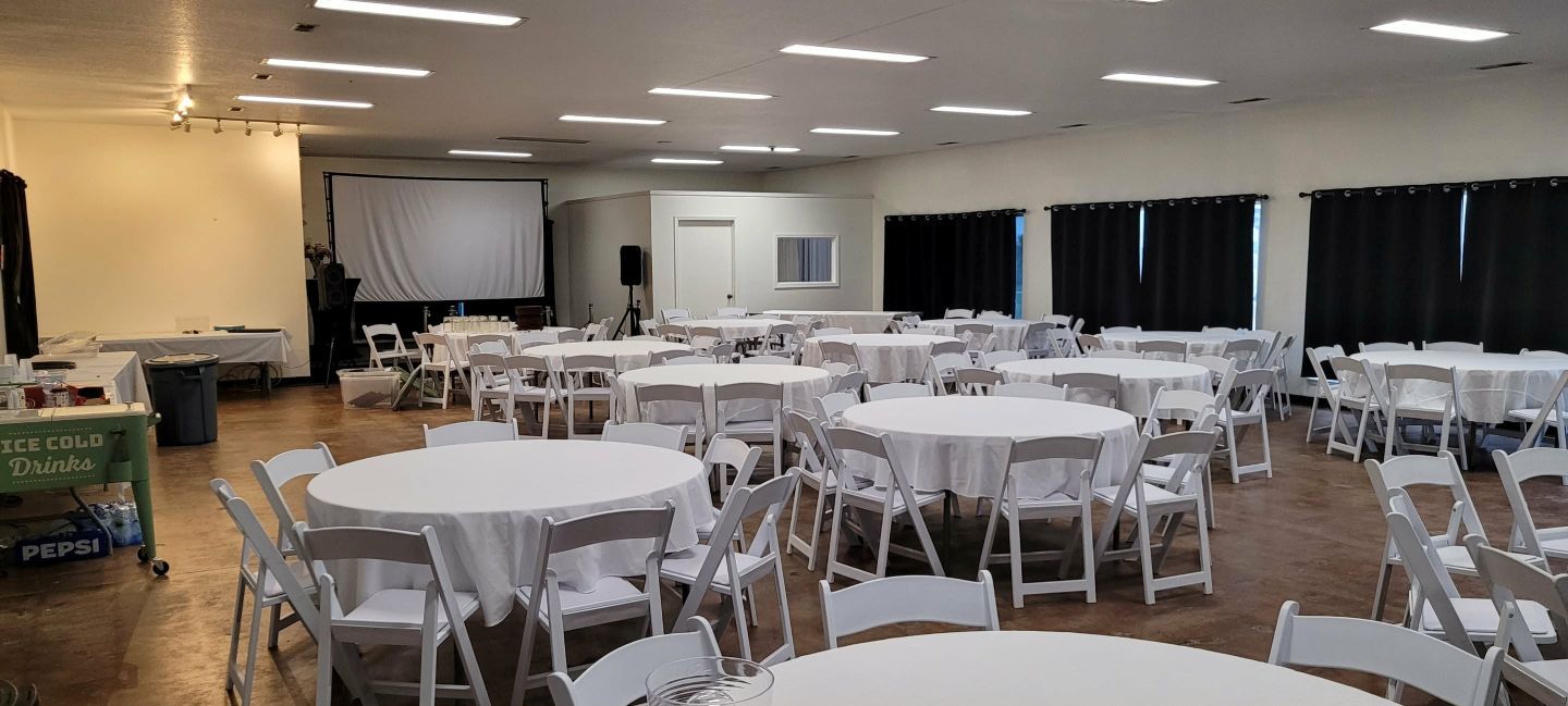 An empty event hall with round tables covered in white tablecloths and white chairs.