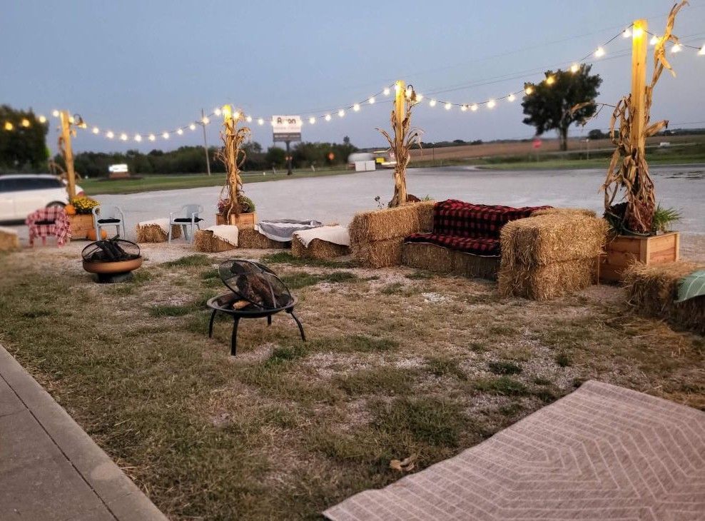 Outdoor seating area with hay bale furniture, string lights, fire pits, and a neutral-colored rug.