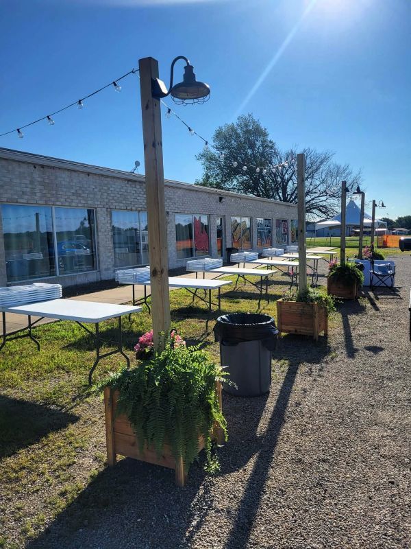 Outdoor event tables set up along a building with string lights and potted plants on a sunny day.