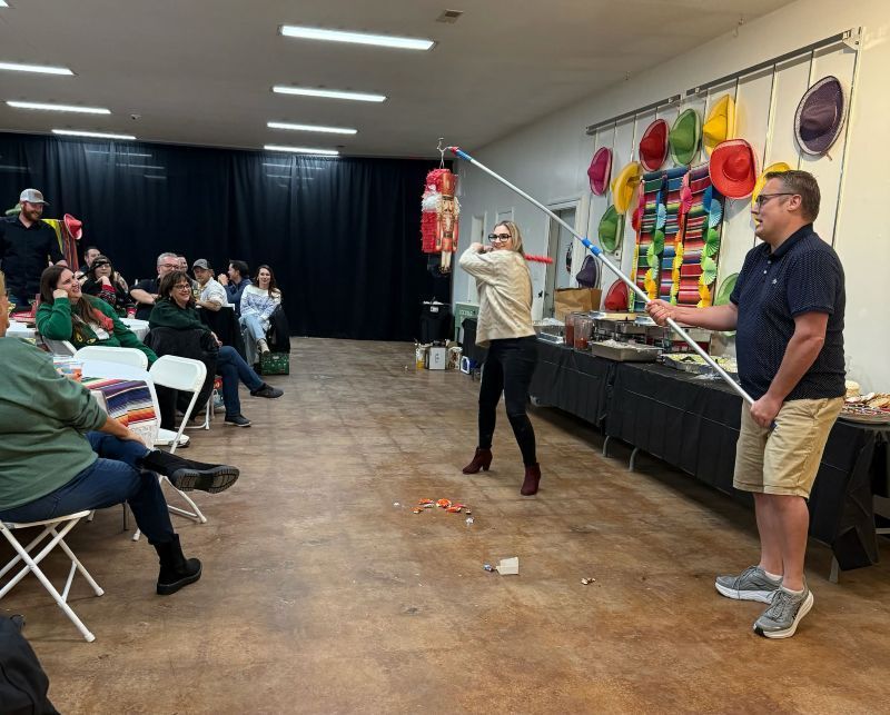 People playing a piñata game at a party. One person swings at the piñata, another guides. Spectators watch.