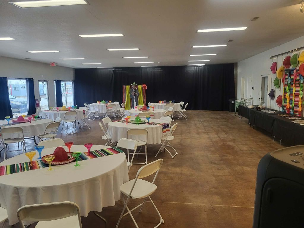 Hall decorated for a party, featuring round tables with white tablecloths, Mexican-themed decor, and a black backdrop.