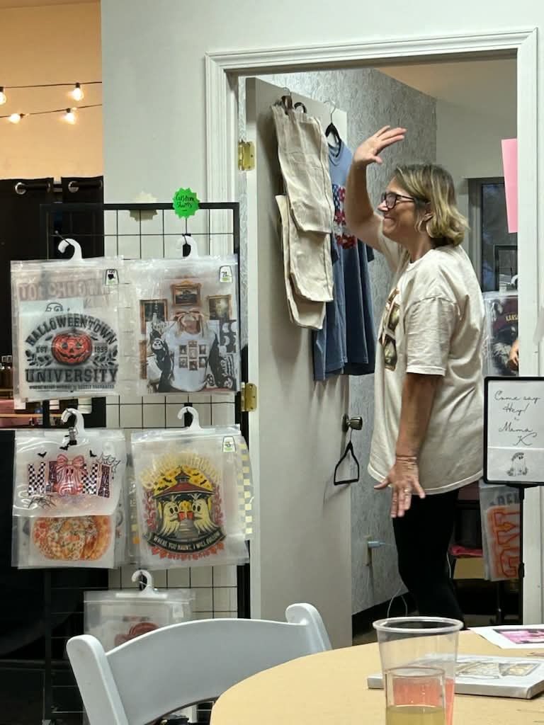 Woman waves from a doorway in a craft fair venue with t-shirts for sale.