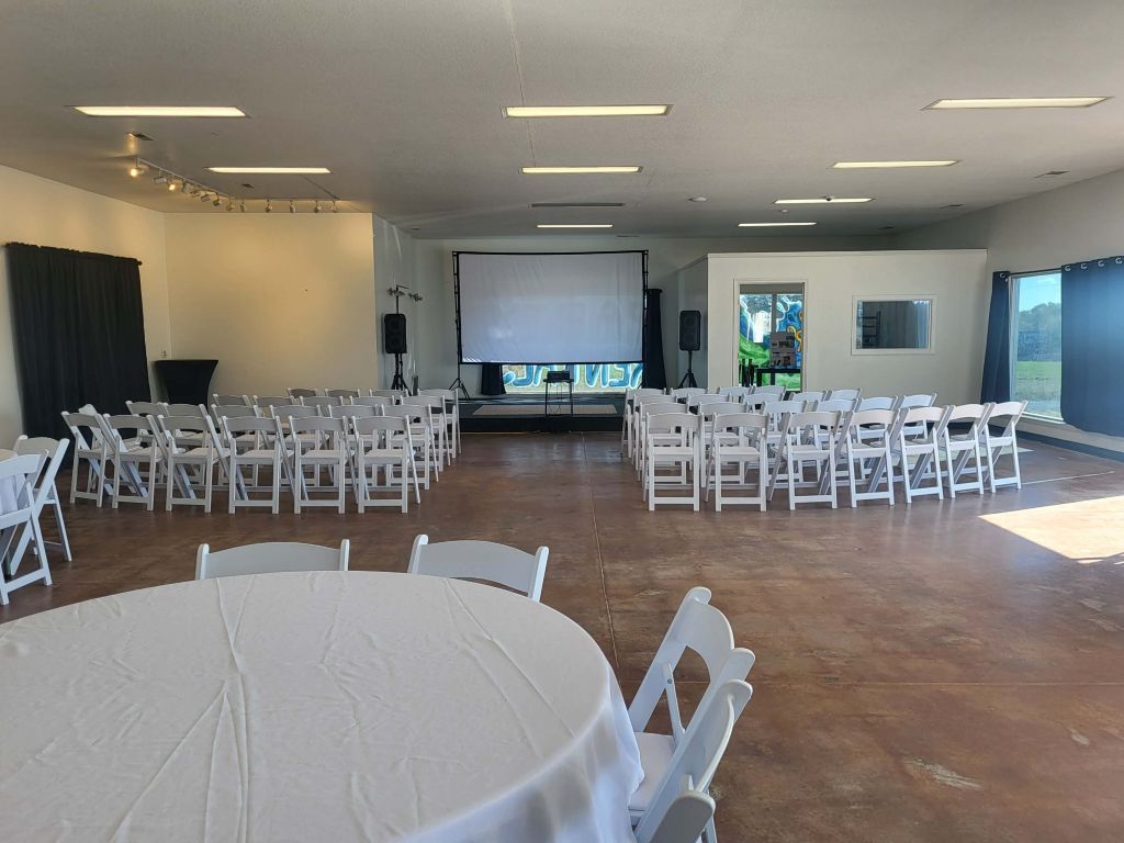 Event space with white chairs and tables, stage with projector screen, and dark curtains.