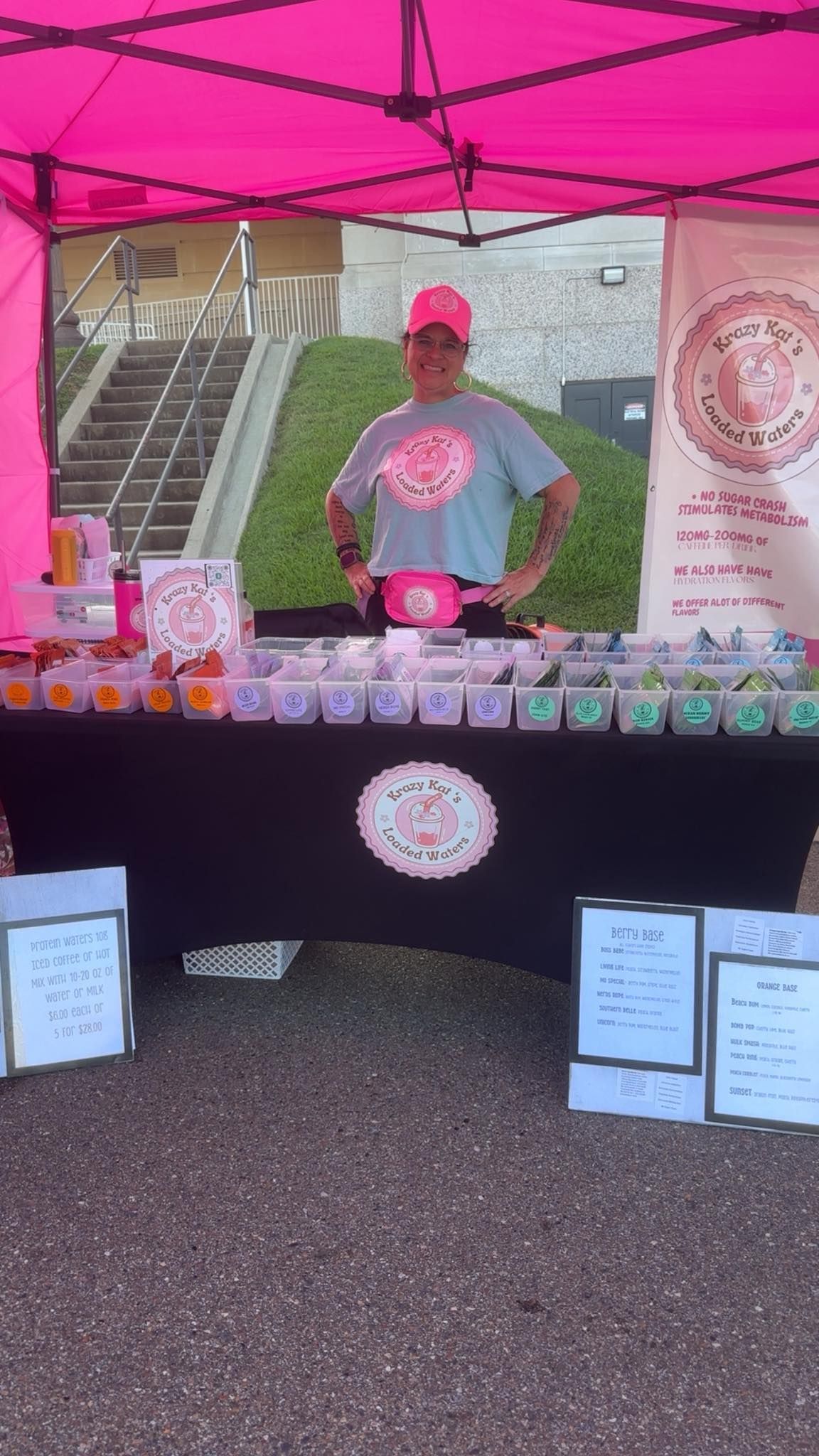 Person standing at a pink-themed vendor booth, with products displayed on a black table.