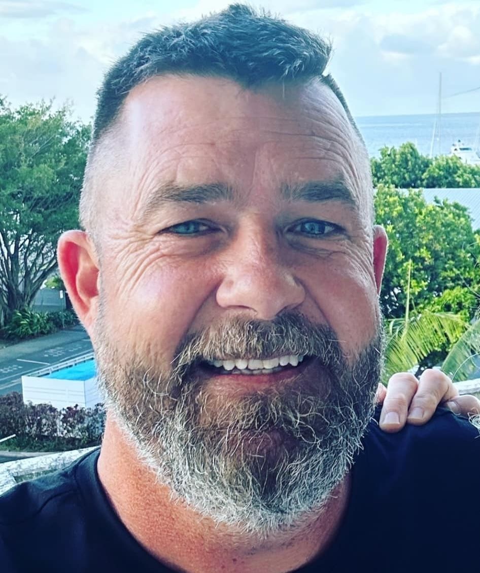 Man with a graying beard, smiling outdoors, wearing a black shirt, trees and ocean in background -Calvin's Mobile Mechanics In Elanora, QLD