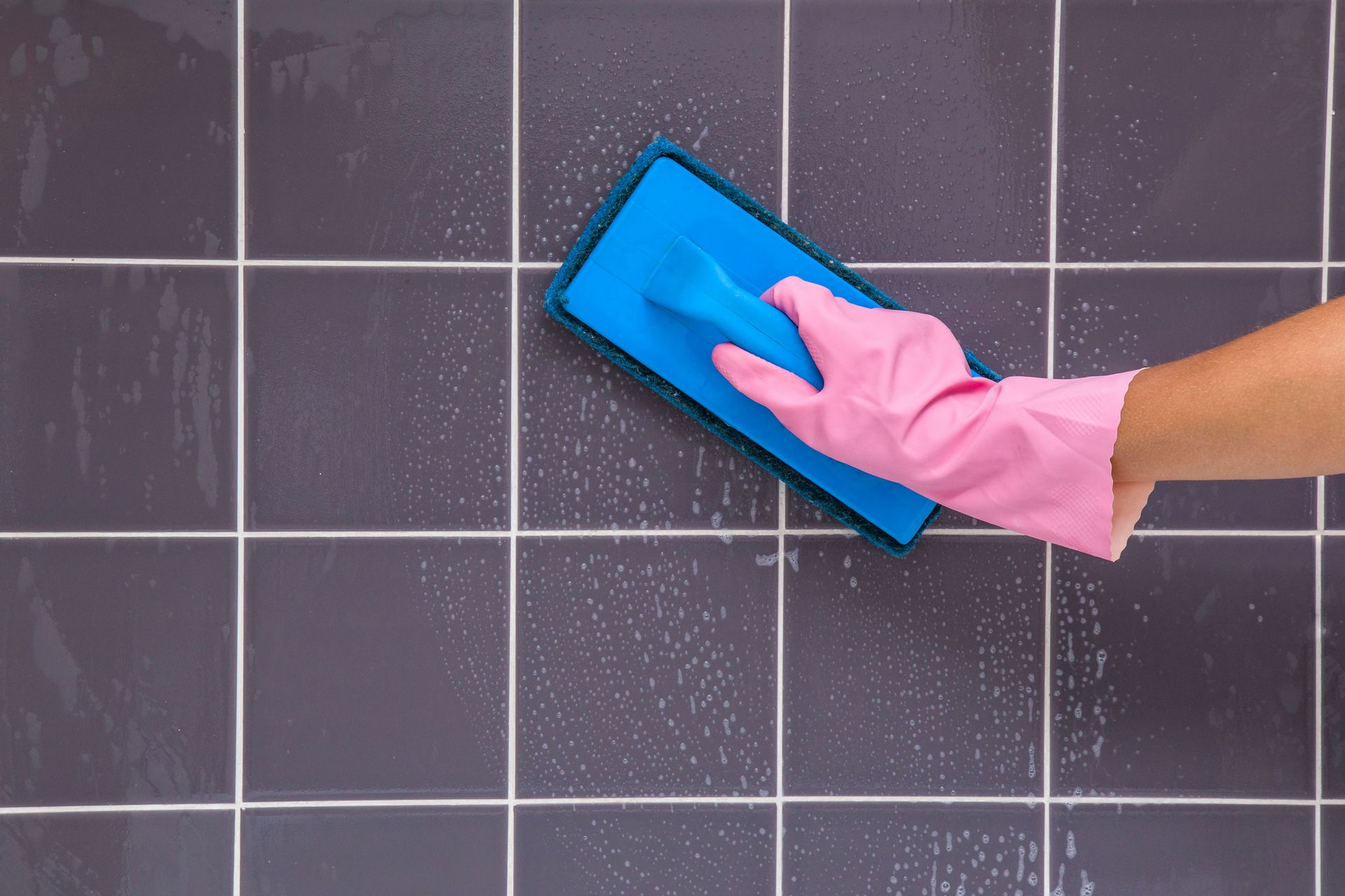 Hand in pink glove scrubbing dark gray tiled wall with blue sponge.