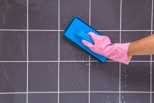 A hand wearing a pink latex glove holds a cleaning pad as it cleans a gray tiled surface.
