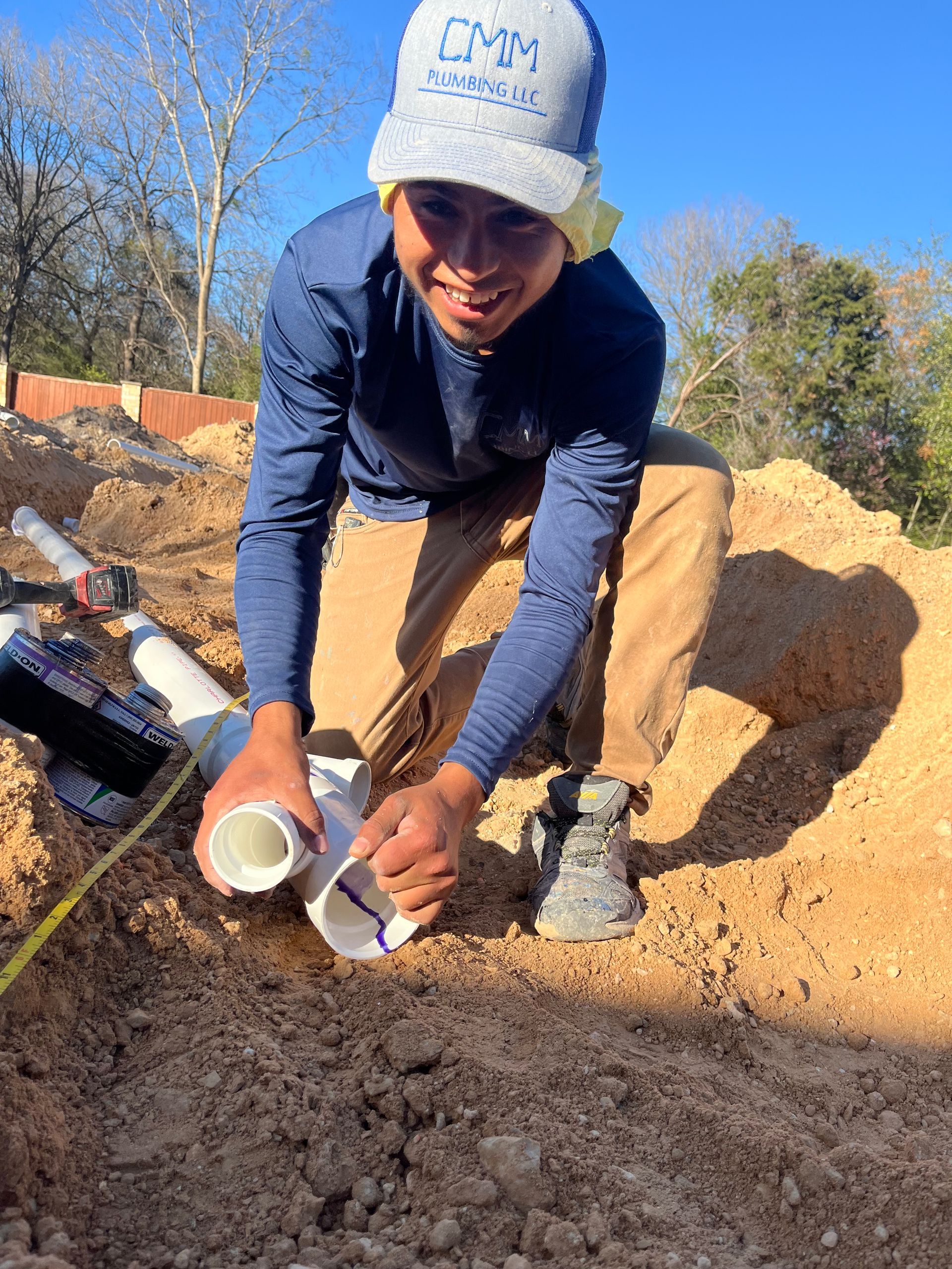 Construction worker fitting PVC pipes in dirt. Wearing a hat, blue shirt, and khaki pants.
