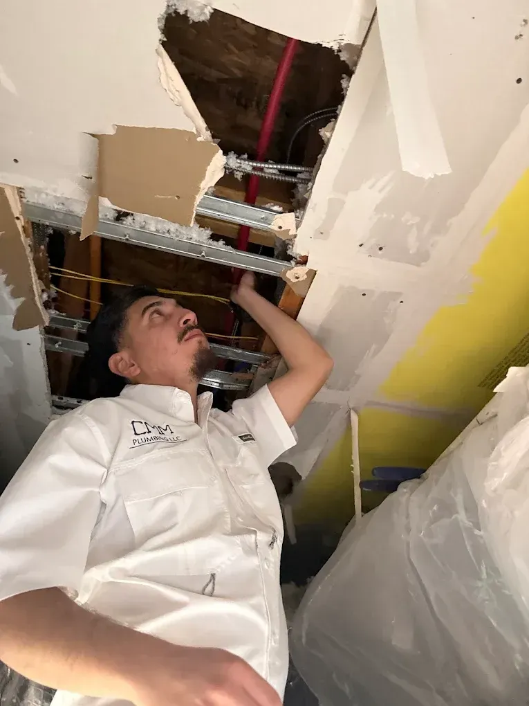 Person in a white coverall inspects a ceiling opening with exposed pipes and wiring