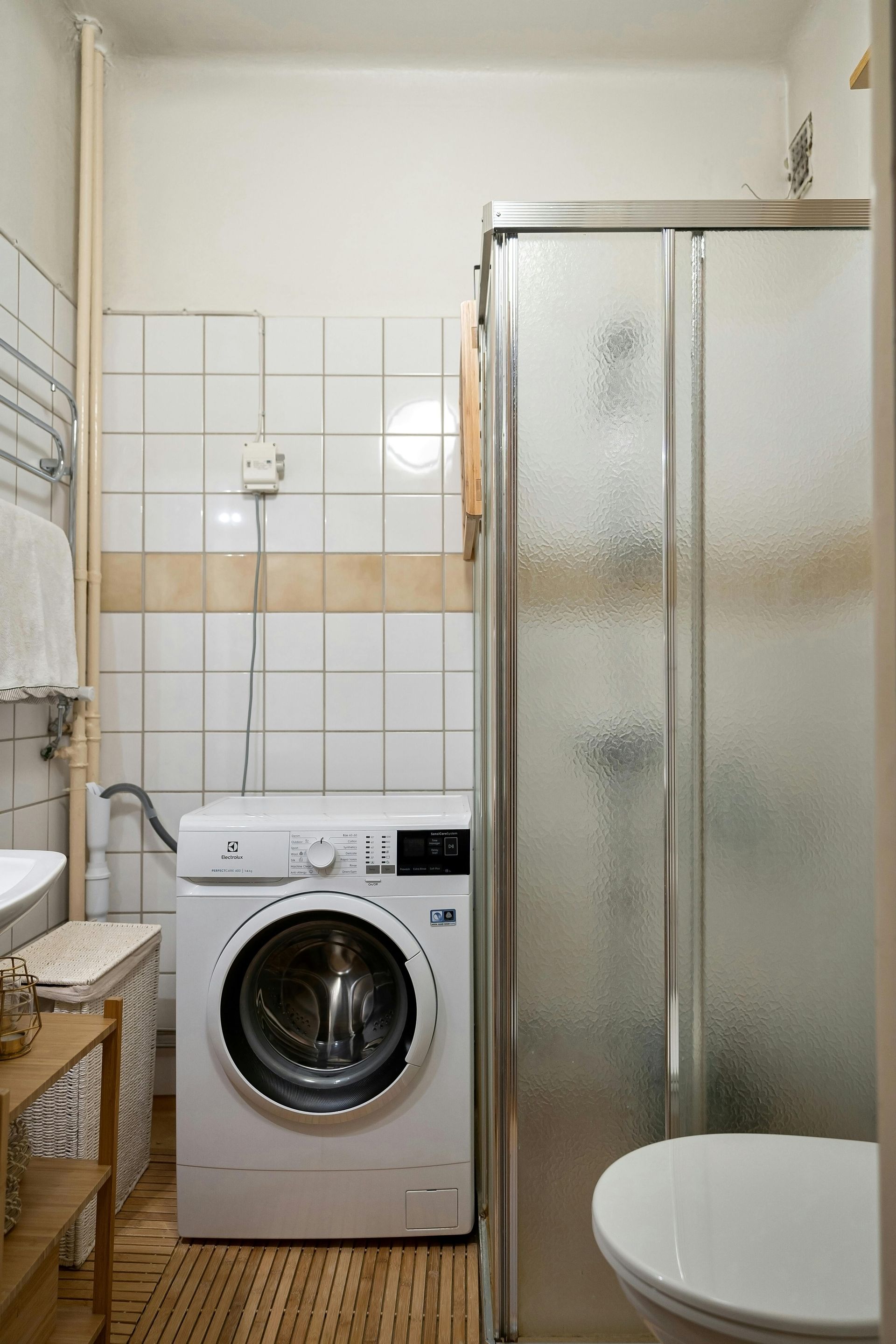 Small bathroom with a washing machine, shower, and toilet. White and beige tile, light wood floor.