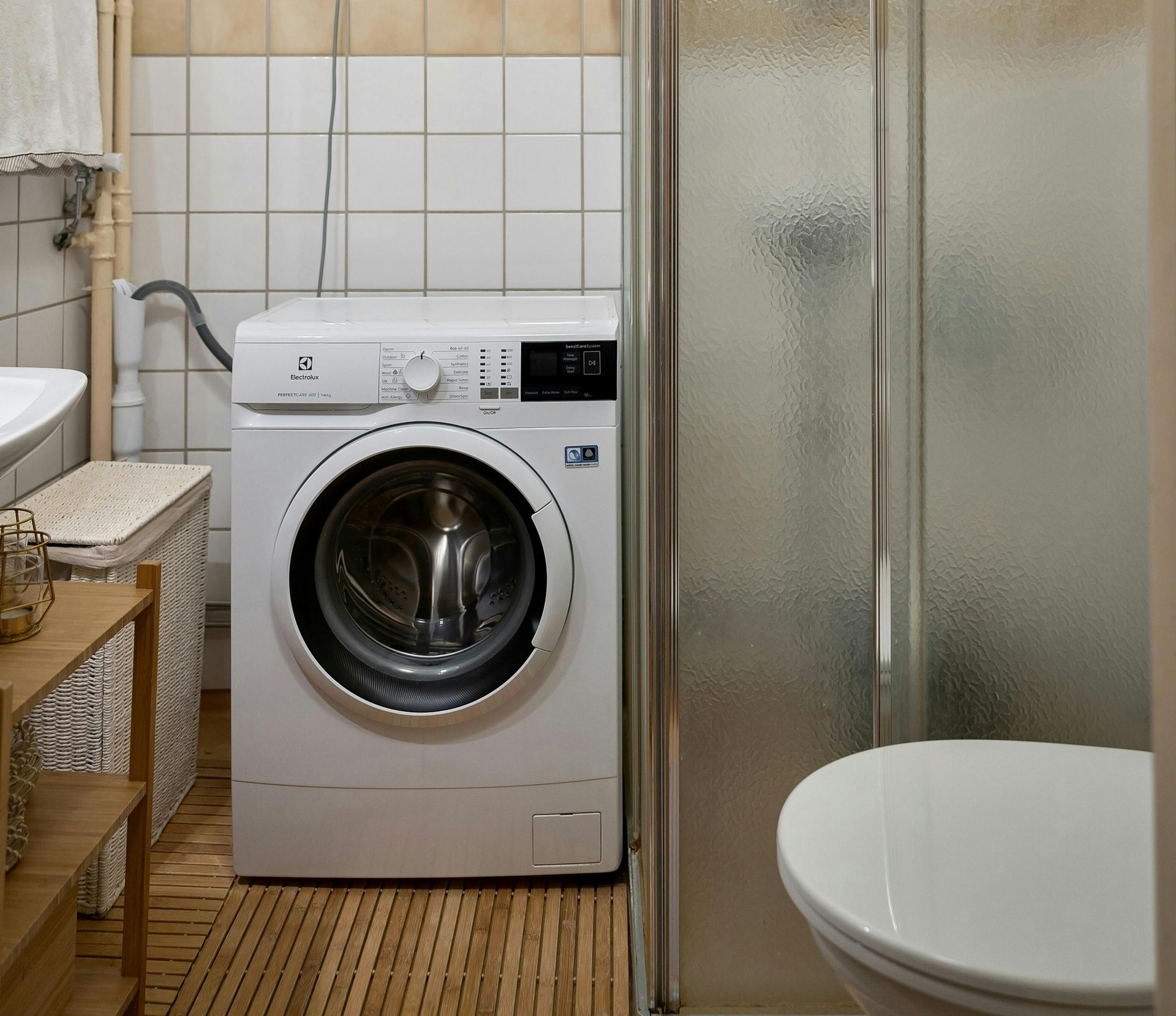A small bathroom with a washing machine, shower, and toilet. Light-colored tiles and wood flooring.