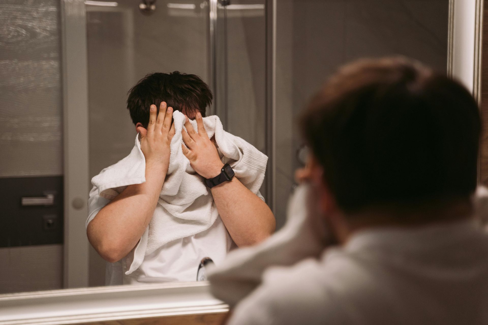 A man is covering his face with a towel in front of a bathroom mirror after showering