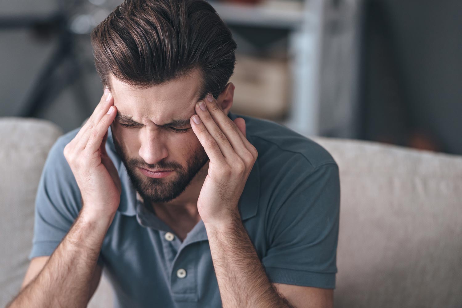 Guy Suffering From Head Ache — McAllen, TX — Texas Rehab Center Man with headache, holding temples, eyes closed, seated on a couch.