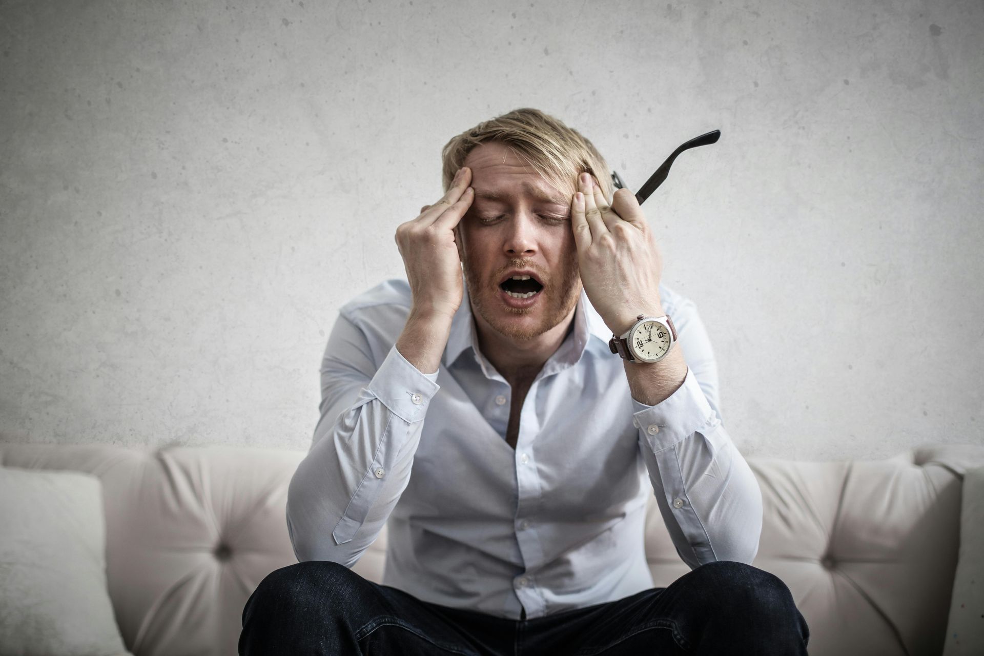 Man with blond hair, wearing a light blue shirt, holding his head in distress on a couch.