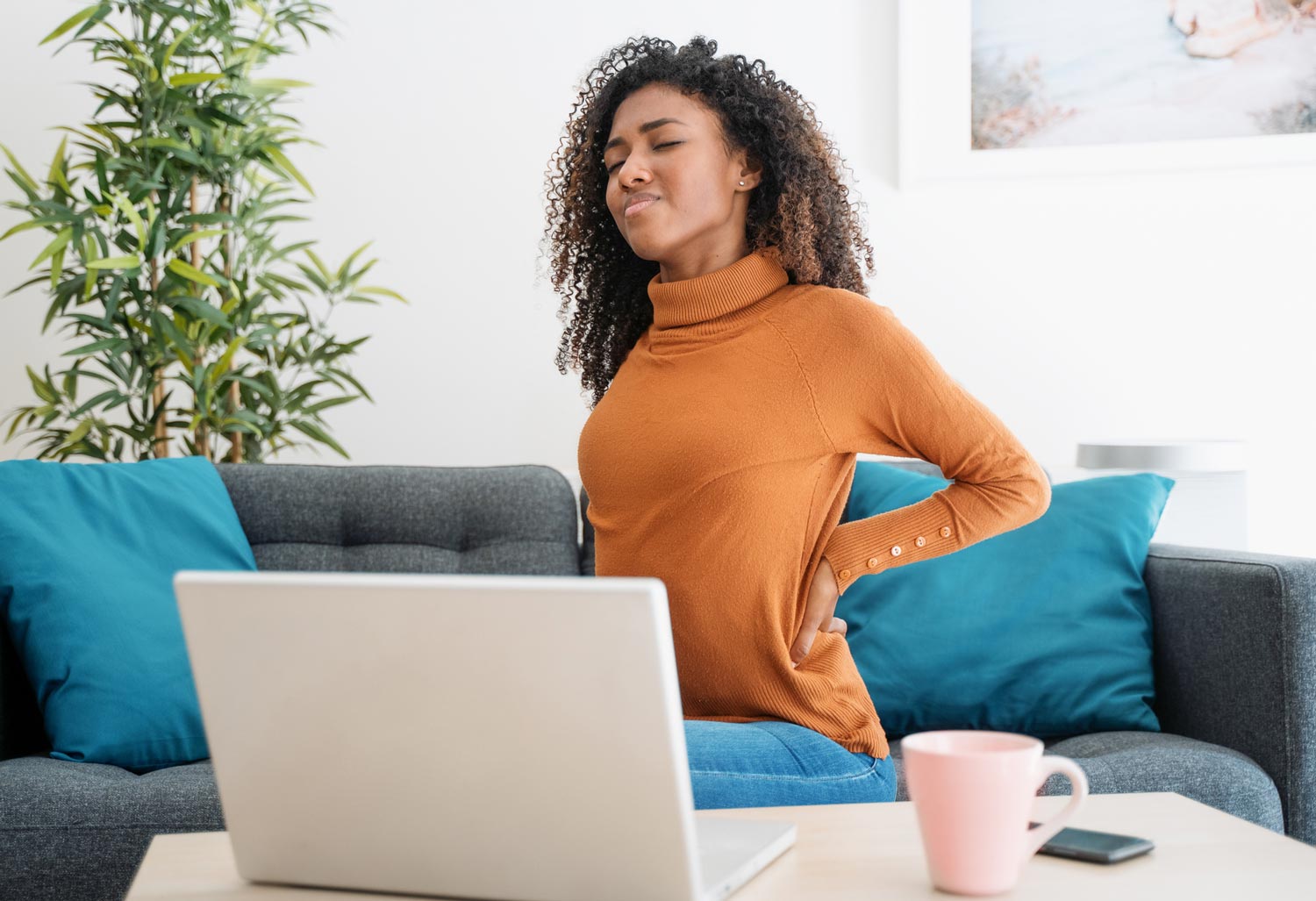 Woman with back pain, seated on a couch with laptop, holding her back.