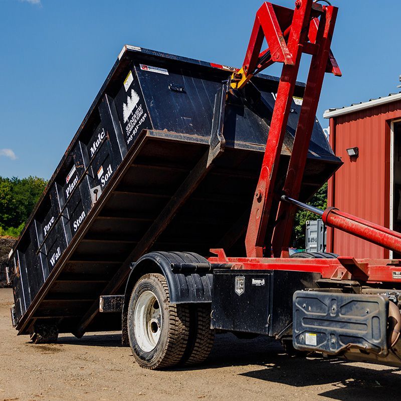 Black dumpster tilted on a red truck, ready to be emptied, in a sunny setting.