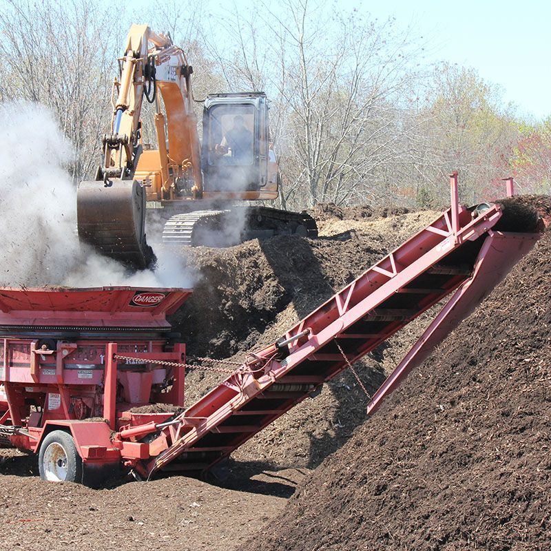 An excavator dumping material into a red conveyor belt, with steam rising.