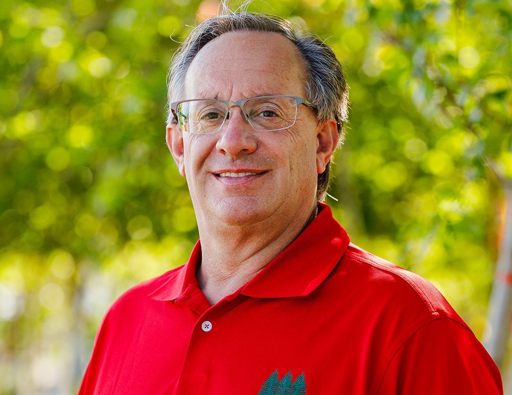 Man with glasses in red polo shirt smiles outdoors, blurred green background.