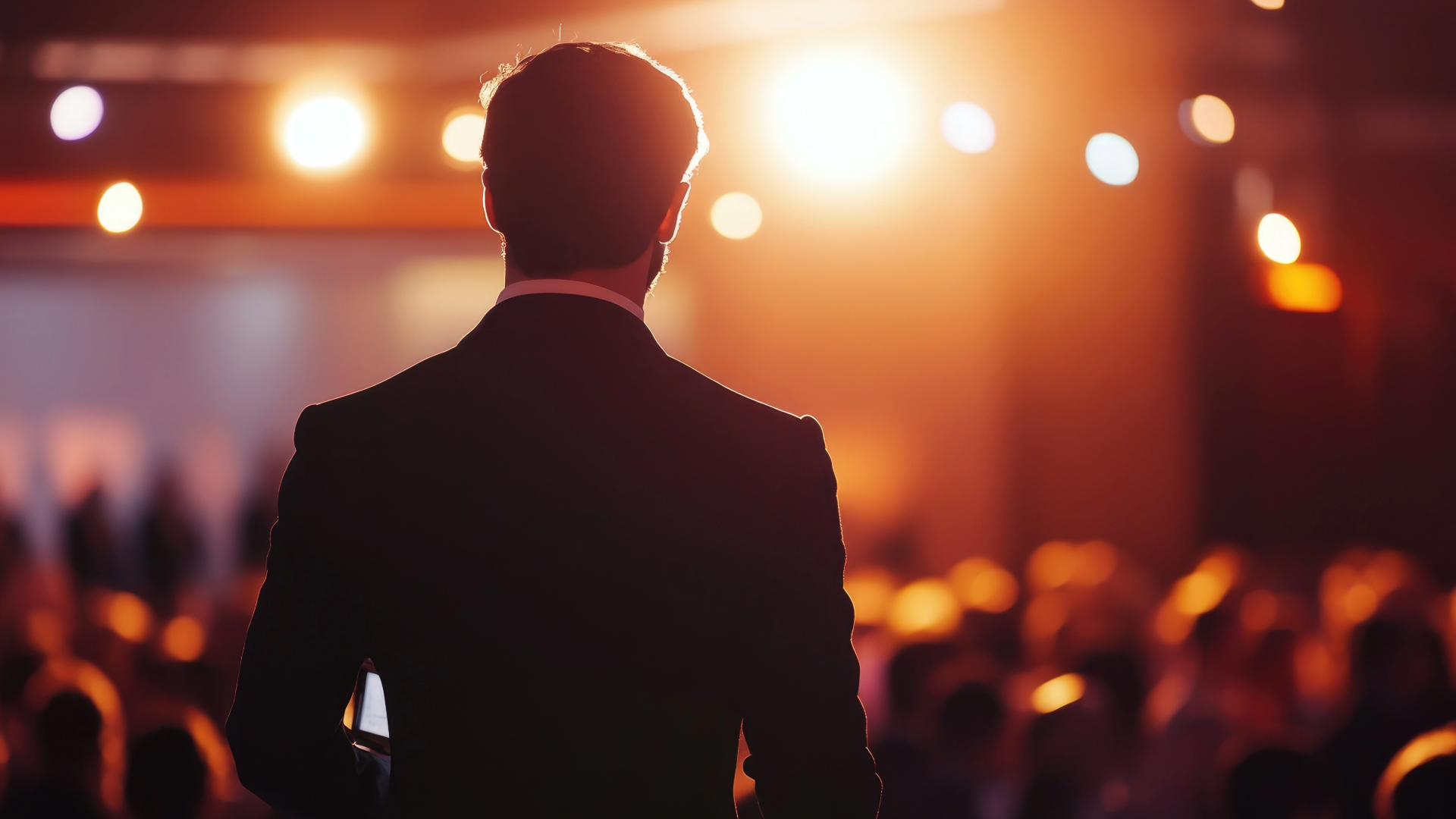 A man in a suit is giving a speech in front of a crowd in a spotlight.