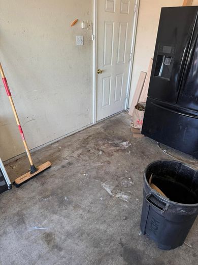 A room with a broom , trash can and refrigerator.