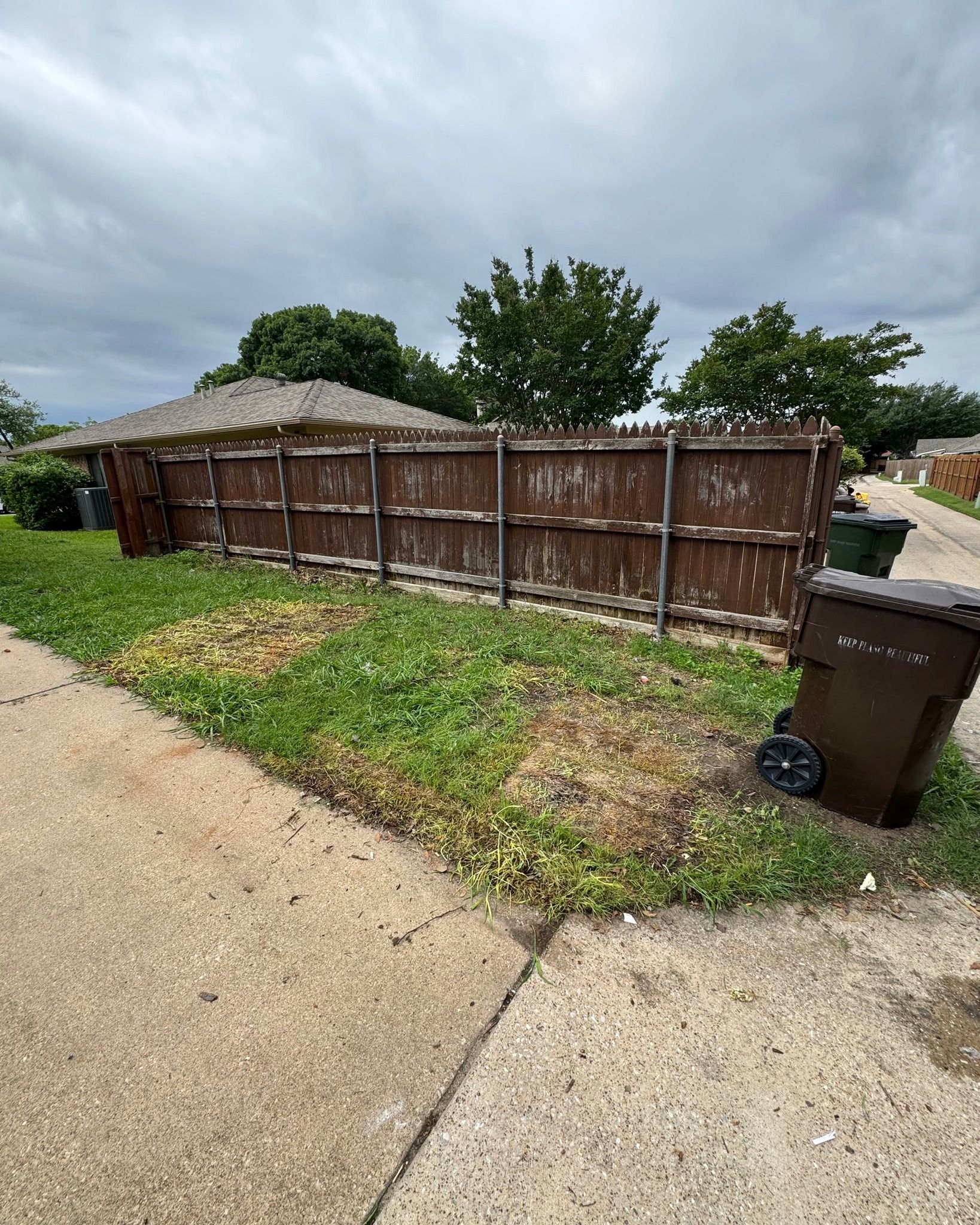 A wooden fence is sitting on the side of a road next to a trash can.