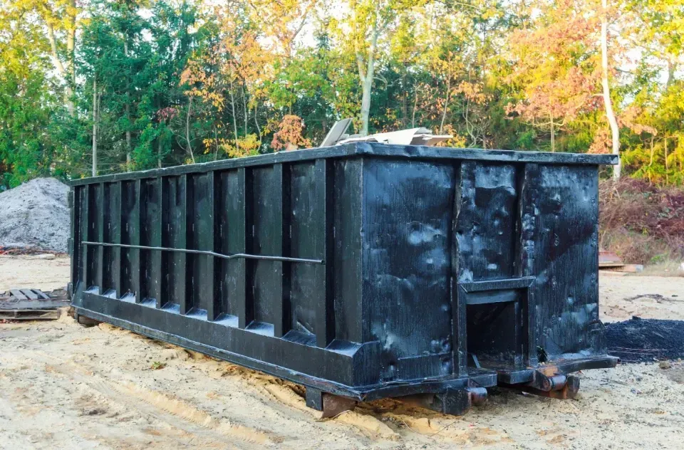 A large black dumpster is sitting in the middle of a dirt field.
