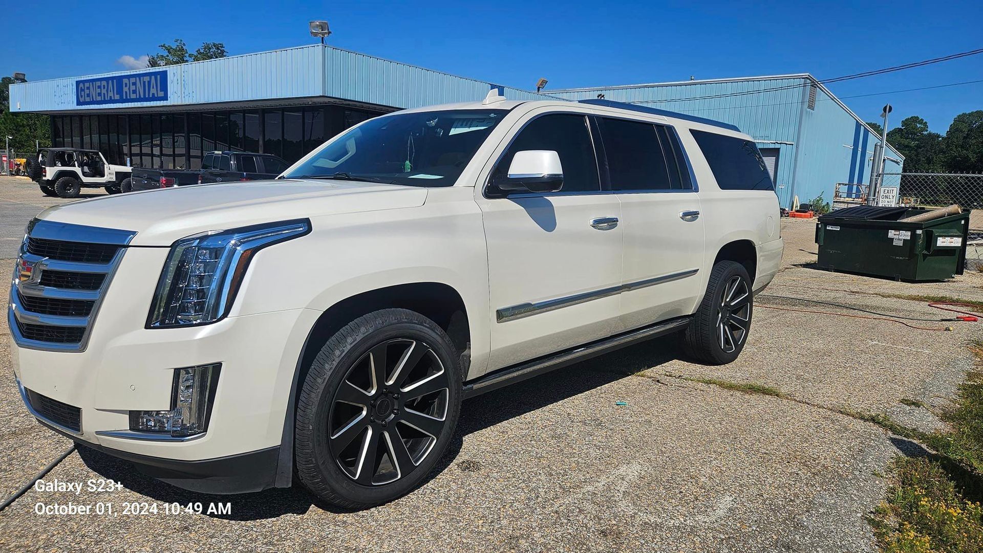 A white cadillac escalade is parked in a gravel lot in front of a building.