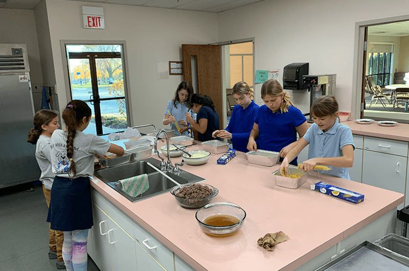 Children in a kitchen, preparing food at a pink counter.  Electives & Clubs | Immanuel Lutheran School | Belvidere, IL